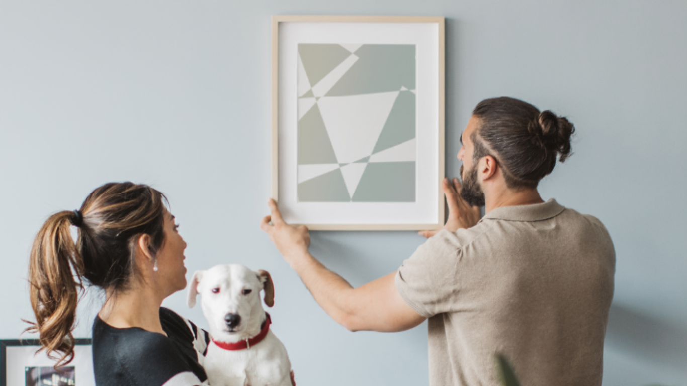 A couple with a dog hanging a picture on a wall