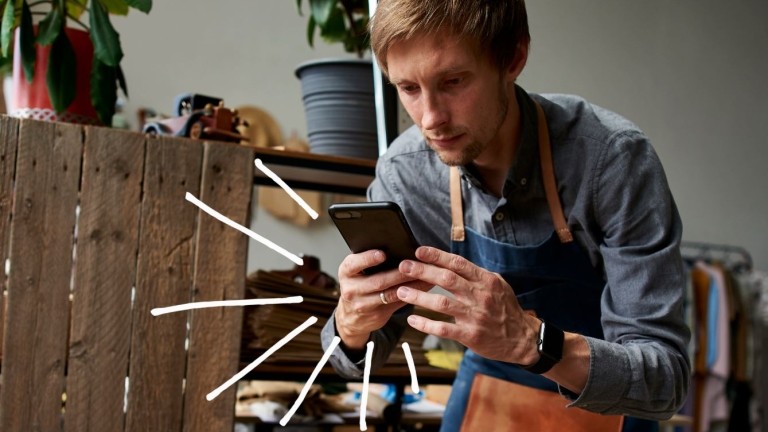Man in shop apron sits in a chair, checking his phone. 