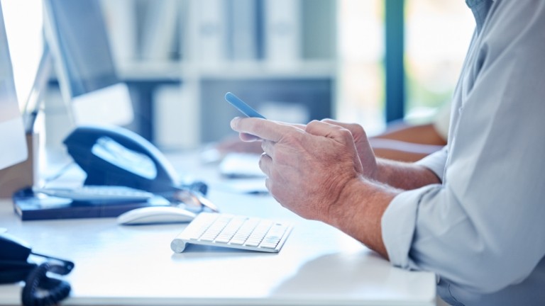 Personâs hands at a desk looking at a mobile phone.