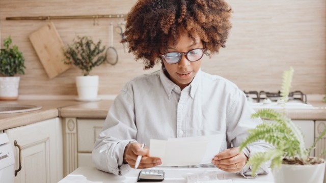 Woman with glasses reviewing finances with calculator at the kitchen table.
