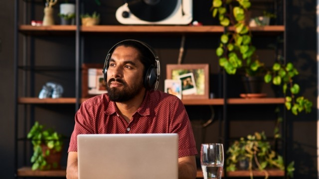 Gentleman sitting in office with laptop and headphones on looking off into the distance.