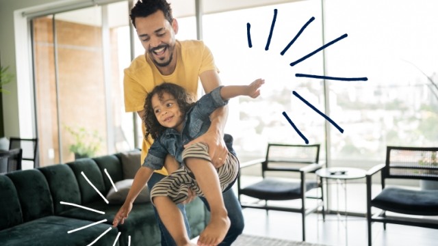Father holding child pretend flying like an airplane in home living room.