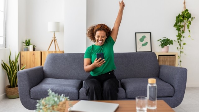 Woman with curly hair in living room on couch with mobile phone and hand raised in celebration.