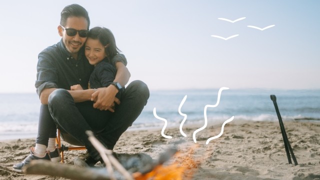 Father and daughter sitting in front of a camp fire on a beach. 