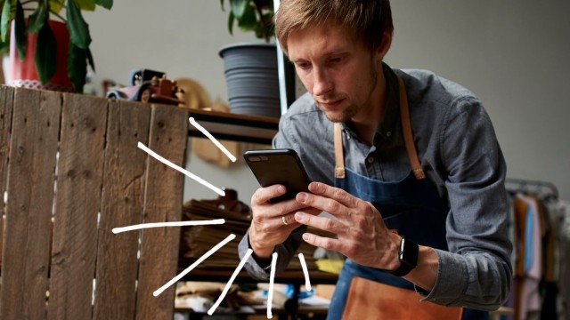 Man in shop apron sits in a chair, checking his phone.