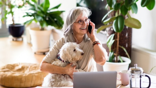 Woman with glasses on the phone and with dog inside a home with plants.