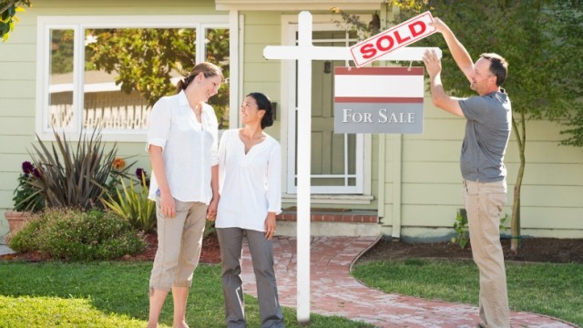 Partners with real estate agent in front of a home with a âSOLDâ sign.