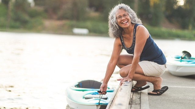Smiling woman with grey hair on a river dock holding onto a kayak.