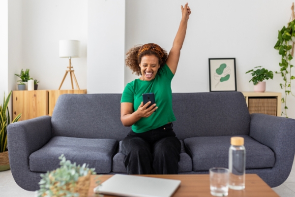 Woman with curly hair in living room on couch with mobile phone and hand raised in celebration.