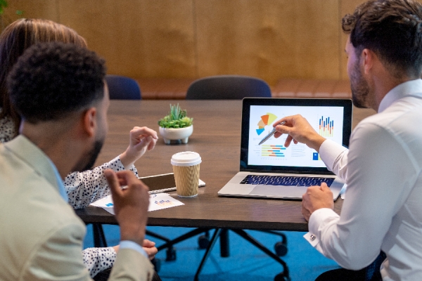 Couple and financial planner sitting at a table reviewing investment performance on a laptop with coffee.    