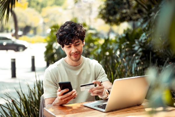 A person sitting outside at a table, checking their computer and phone with a credit card in-hand. 