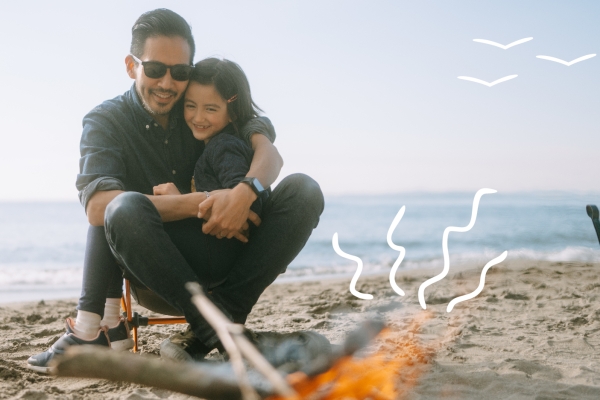 Father and daughter sitting in front of a camp fire on a beach. 