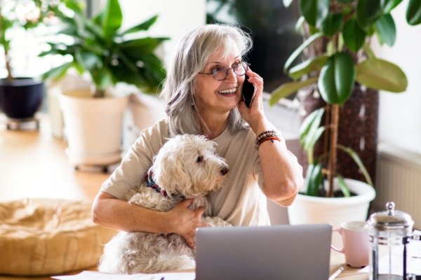 Woman with glasses on the phone and with dog inside a home with plants.