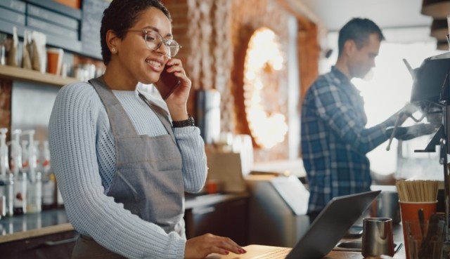 Coffee shop owner in apron and glasses working on a laptop and having a conversation on a mobile phone.